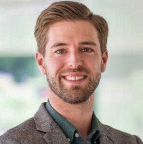 A man with light brown hair and a trimmed beard, wearing a grey blazer and green shirt, smiles at the camera against a blurred background.