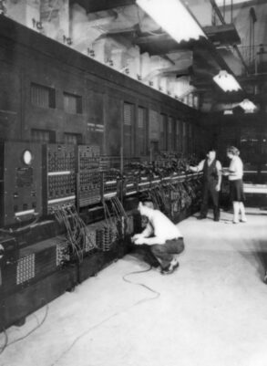 Several people operate a large room-sized early computer with numerous cables and panels, likely from the mid-20th century.