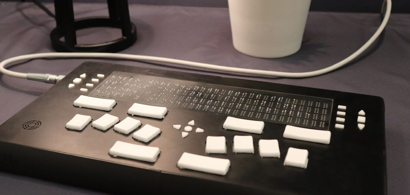 A refreshable braille display with white keys sits on a table near a white potted plant and a lamp.