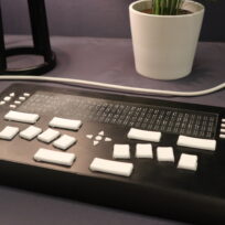 A refreshable braille display with white keys sits on a table near a white potted plant and a lamp.