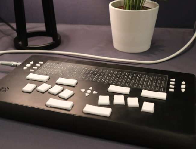 A refreshable braille display with white keys sits on a table near a white potted plant and a lamp.