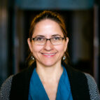 A woman with glasses and brown hair tied back smiles at the camera. She is wearing a blue shirt and dark sweater, standing indoors with a blurred background.