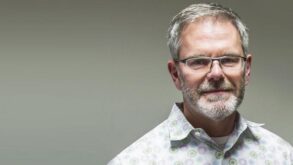 A middle-aged man with gray hair, glasses, and a beard is wearing a patterned shirt and standing against a plain, light background.