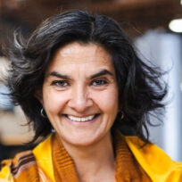 Woman with dark hair and a yellow scarf smiles at the camera in an indoor setting with a blurred background.