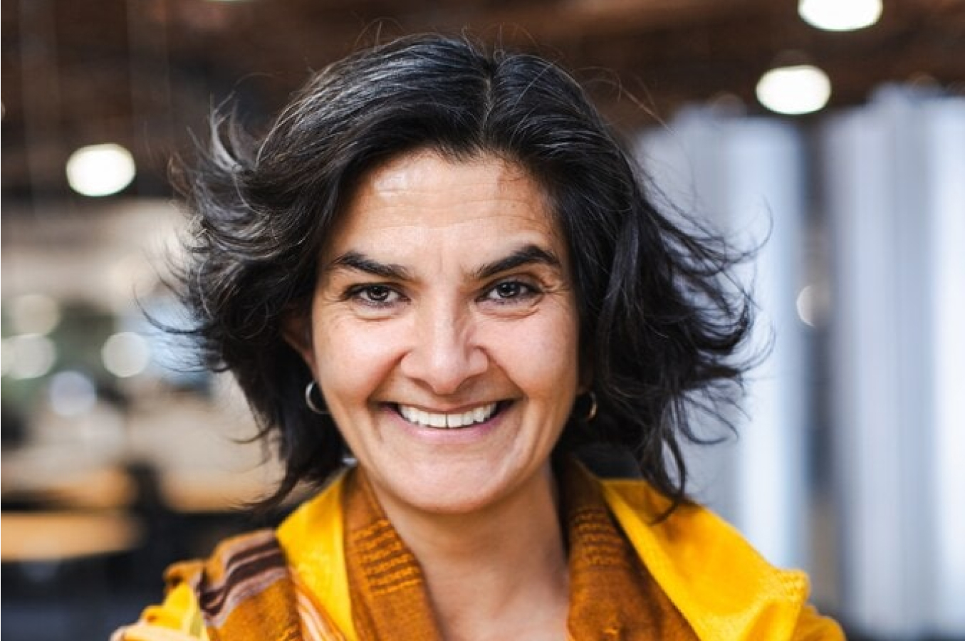 Woman with dark hair and a yellow scarf smiles at the camera in an indoor setting with a blurred background.