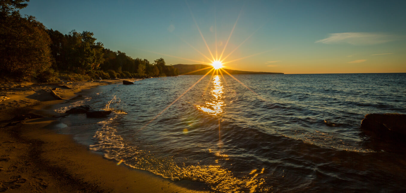 The sun sets over a calm lake, casting a bright reflection on the water near a sandy, tree-lined shoreline.