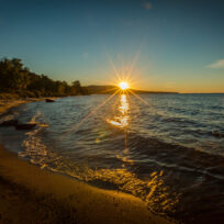 The sun sets over a calm lake, casting a bright reflection on the water near a sandy, tree-lined shoreline.