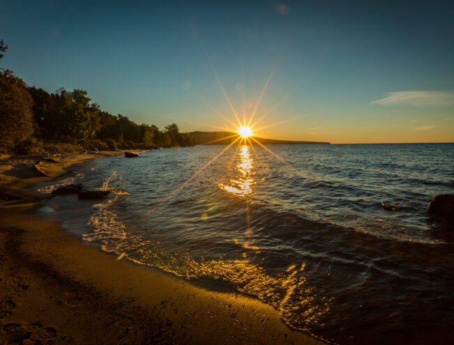 The sun sets over a calm lake, casting a bright reflection on the water near a sandy, tree-lined shoreline.