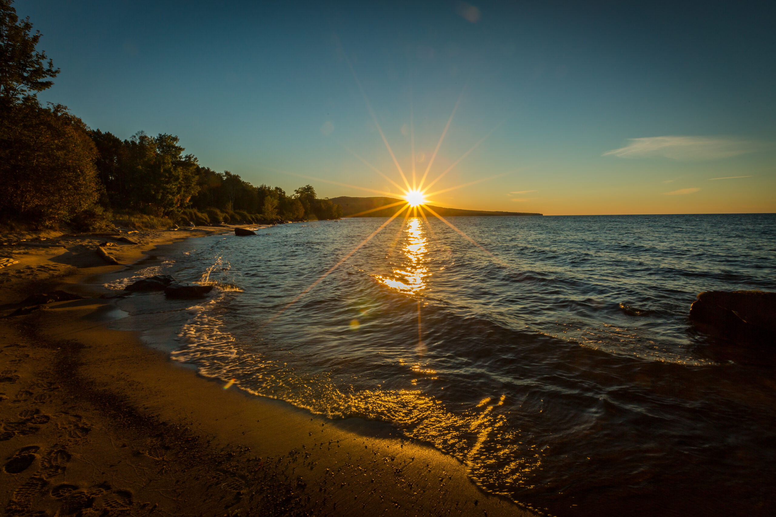 The sun sets over a calm lake, casting a bright reflection on the water near a sandy, tree-lined shoreline.