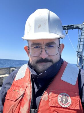 Person wearing a white hard hat, glasses, and an orange "Mustang Survival" life vest stands on a boat with the ocean and blue sky in the background.