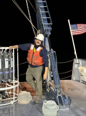 A person in a hard hat and orange life vest stands on a ship's deck at night near scientific equipment, with the US flag visible in the background.