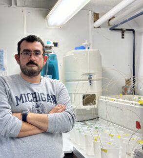 A person wearing a Michigan sweatshirt stands with arms crossed in a laboratory, surrounded by scientific equipment, plastic cups, and tubes.