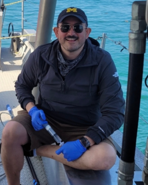 A man wearing a cap, sunglasses, and gloves sits on a boat while holding a syringe-like tool near his bare foot over turquoise water.