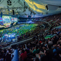 Large crowd watches an esports tournament in an indoor arena, with a brightly lit stage and multiple large screens displaying the game.