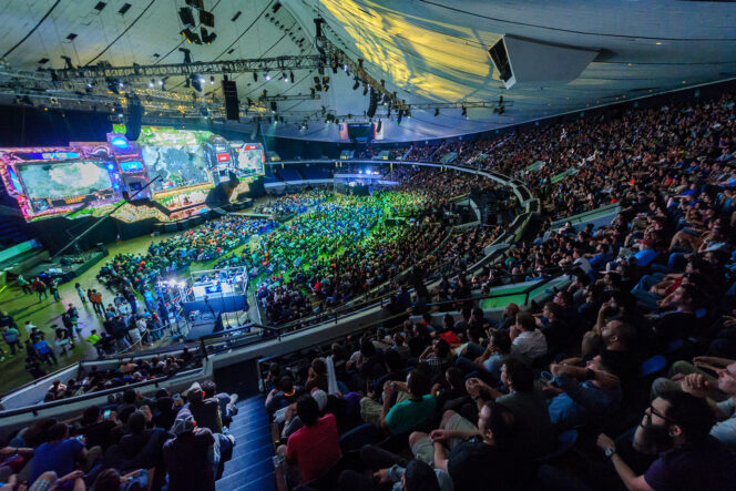 Large crowd watches an esports tournament in an indoor arena, with a brightly lit stage and multiple large screens displaying the game.