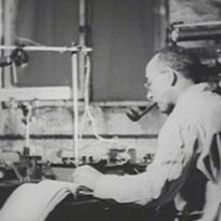A man wearing glasses and a lab coat smokes a pipe while working with scientific equipment and taking notes at a laboratory desk.