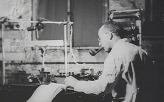 A man wearing glasses and a lab coat smokes a pipe while working with scientific equipment and taking notes at a laboratory desk.