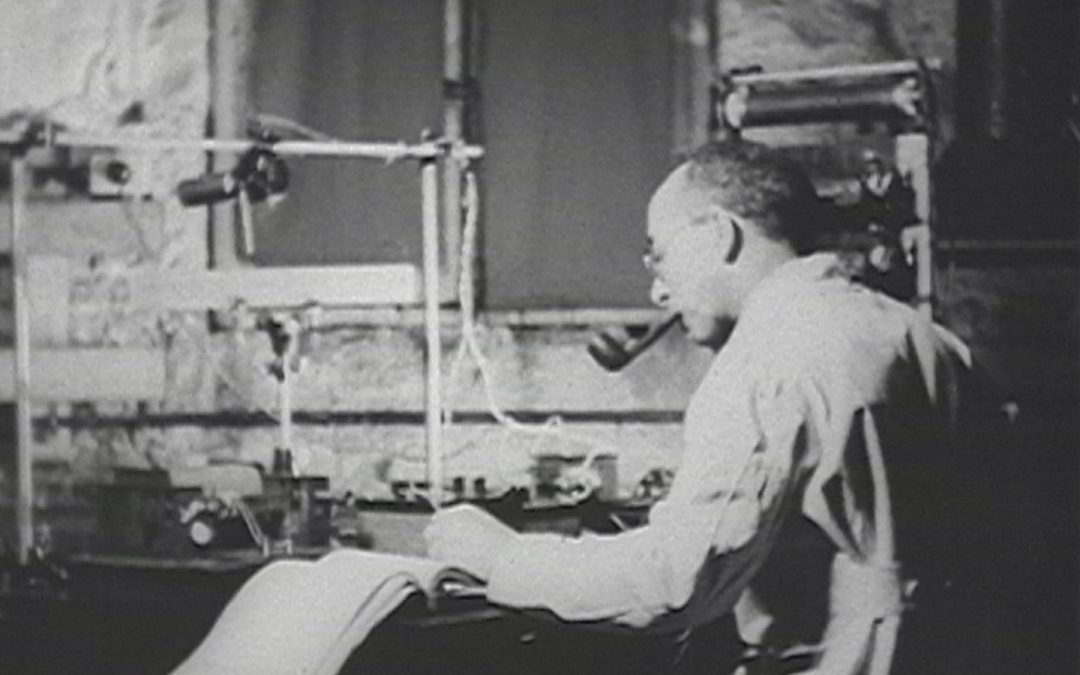 A man wearing glasses and a lab coat smokes a pipe while working with scientific equipment and taking notes at a laboratory desk.