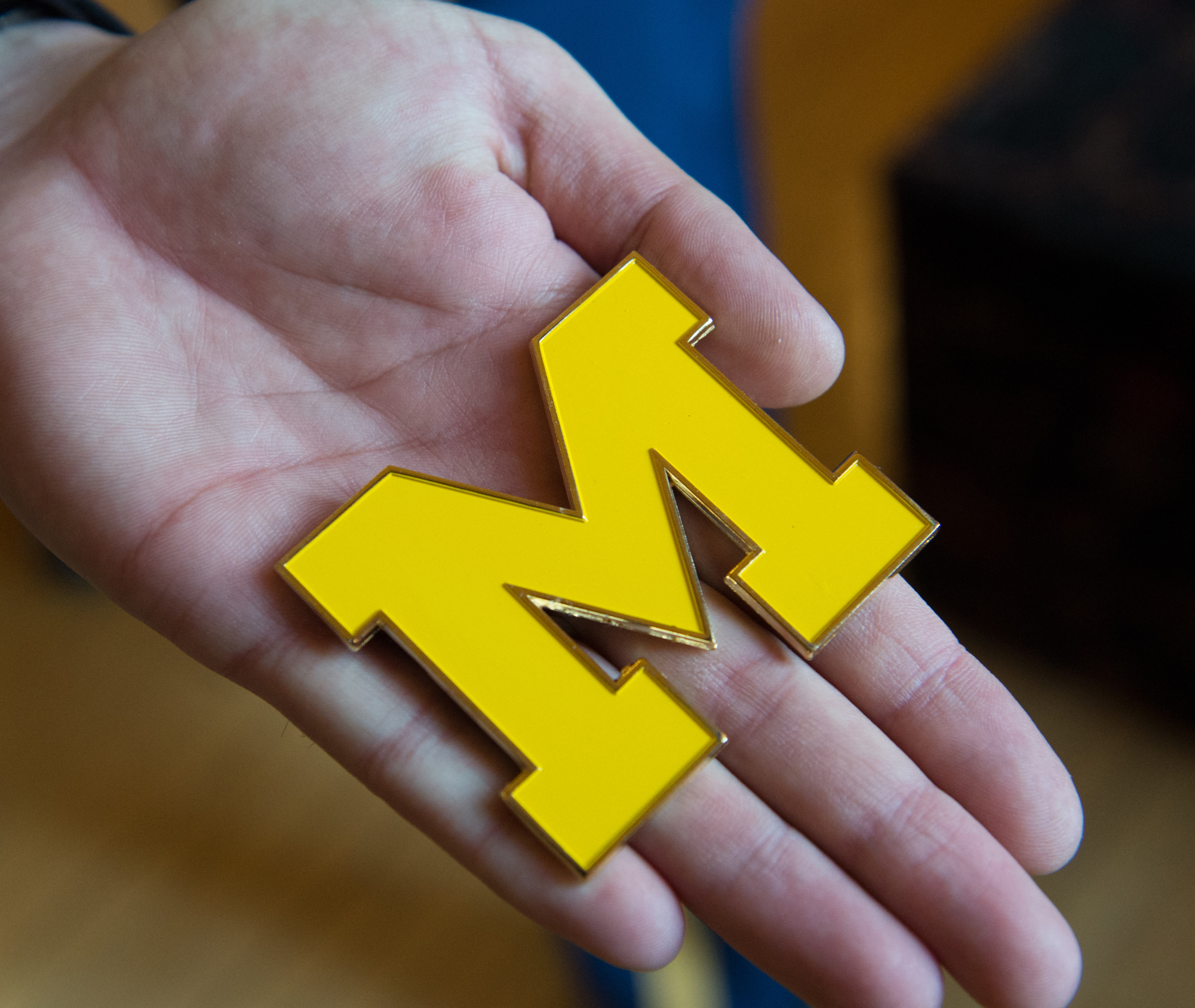 A person holds a large, yellow, block letter "M" pin or emblem in their open palm.