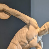 Marble statue of a male athlete preparing to throw a discus, set against a plain blue-gray background.