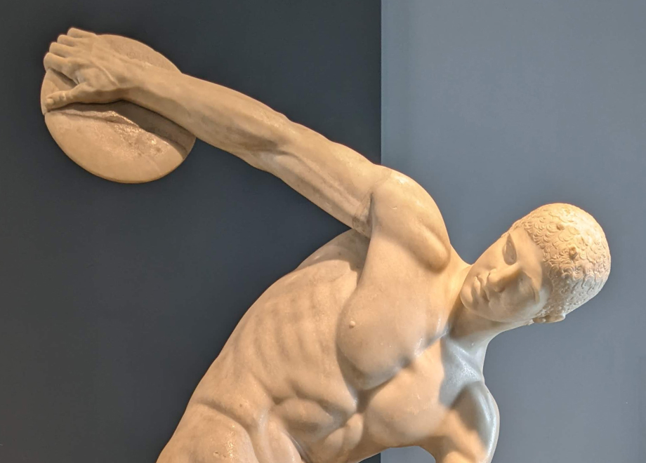 Marble statue of a male athlete preparing to throw a discus, set against a plain blue-gray background.
