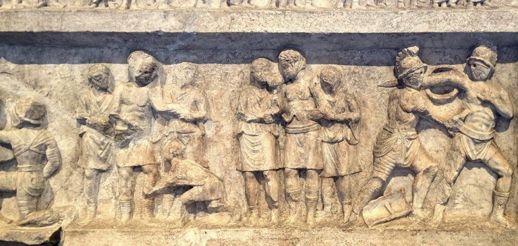 Ancient stone relief shows multiple figures standing, interacting, and two men engaged in combat with shields and helmets on the right side.