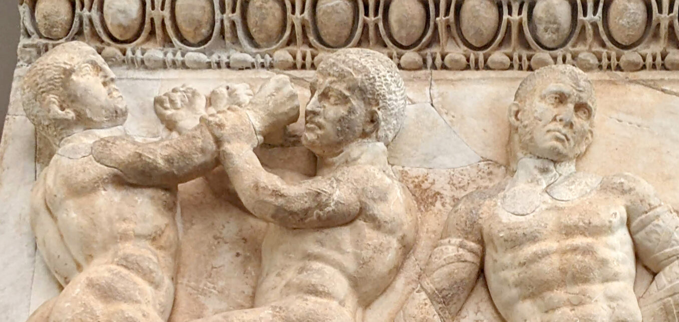Ancient stone relief depicting two muscular men wrestling and a third man watching, with decorative patterns above them.