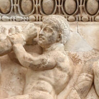 Ancient stone relief depicting two muscular men wrestling and a third man watching, with decorative patterns above them.