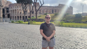 A person stands on a cobblestone plaza in front of the Colosseum in Rome, wearing sunglasses, a black shirt, and shorts, with sunlight streaming over them.
