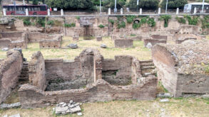 Ancient brick ruins with partial walls and open foundations, surrounded by grass, near a road with a red bus and people in the background.