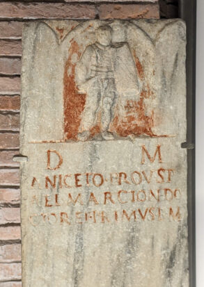 Ancient stone tablet with a carved image of a robed figure above a Latin inscription; the tablet is displayed against a brick wall.