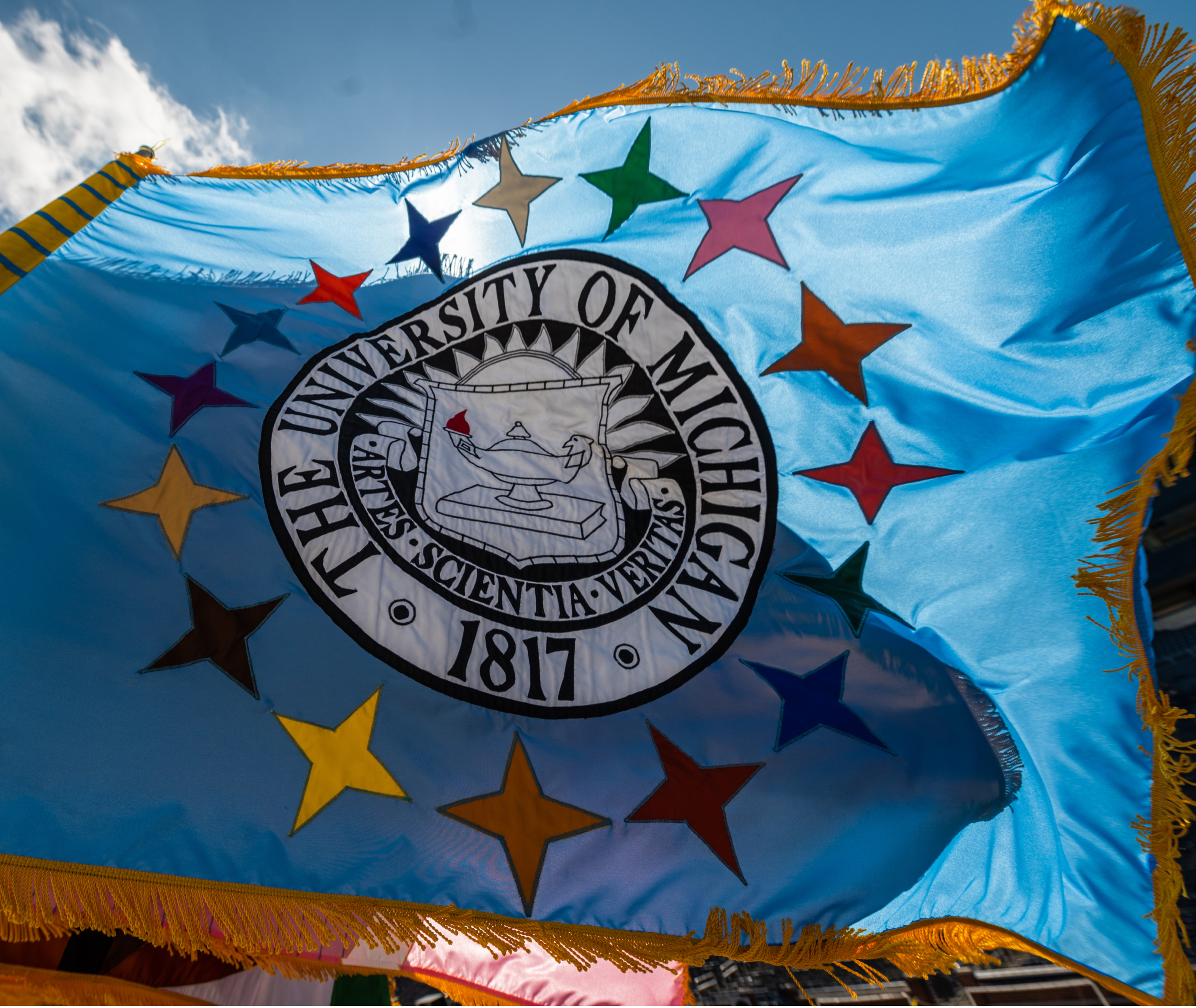 A blue University of Michigan flag with multicolored stars and the university seal flutters in the wind under a partly cloudy sky.