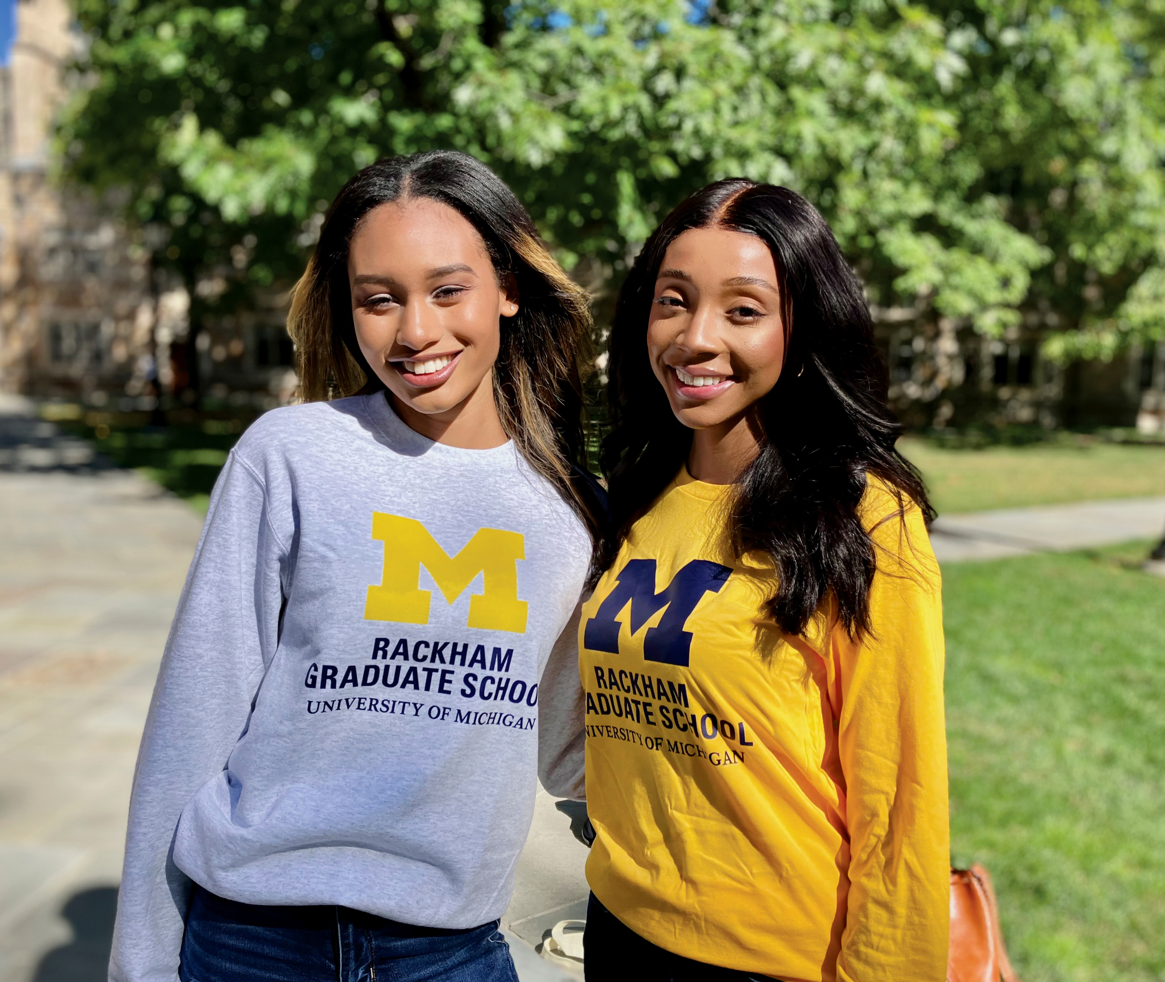 Two women standing outdoors, smiling, wearing University of Michigan Rackham Graduate School sweatshirts, one in grey and one in yellow, with trees and a building in the background.