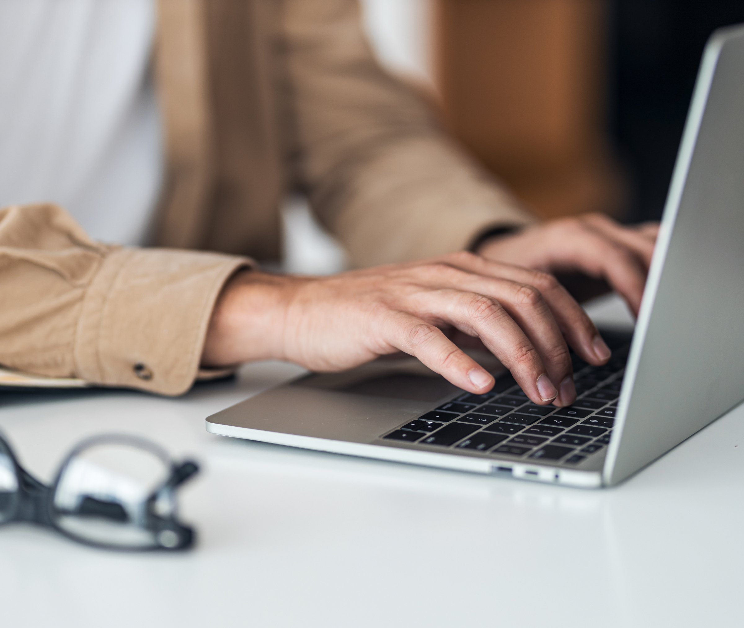 A person types on a laptop at a desk with a pair of eyeglasses nearby.