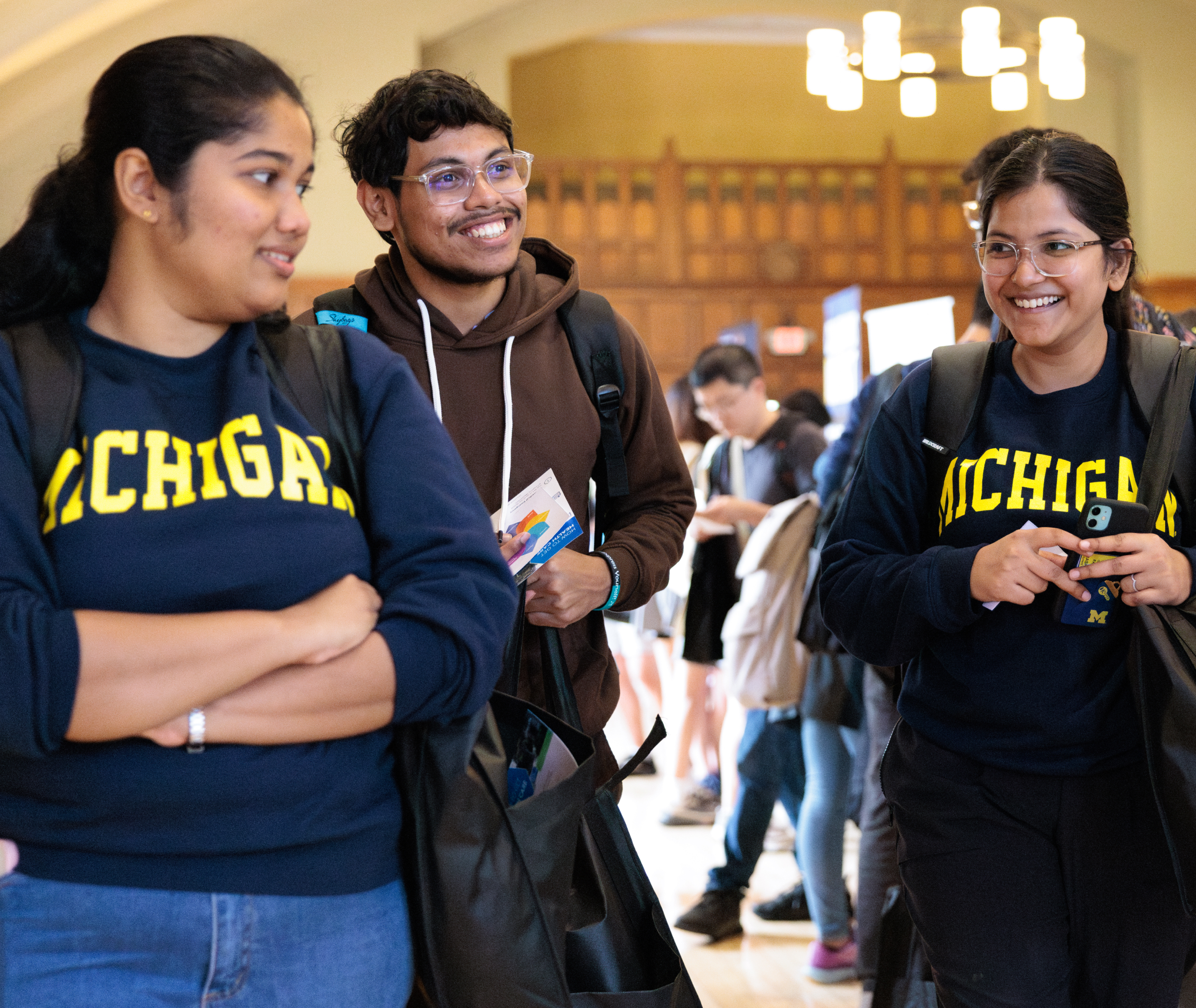 Three students wearing University of Michigan shirts stand indoors, smiling and talking, with other people visible in the background.