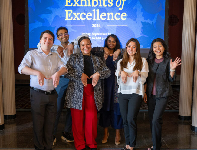 Six people stand in front of a screen displaying "Exhibits of Excellence 2024," all posing with their hands bent downward, smiling at the camera.
