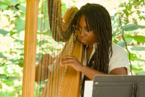 A person with long dreadlocks and glasses plays a wooden harp outdoors, with green foliage in the background and sheet music in front of them.