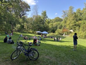 People sit and relax on a grassy field near trees, with bicycles in the foreground and a small outdoor stage and umbrellas in the background under a clear sky.