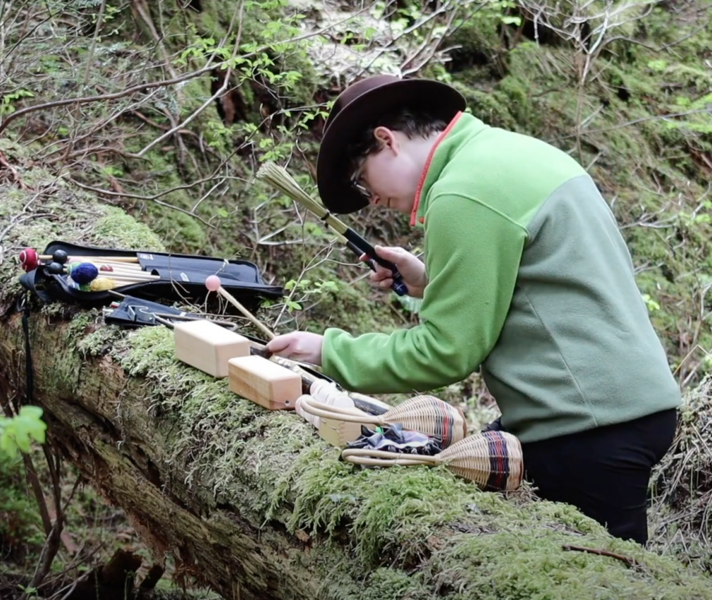 Alexis Lamb uses a handheld recorder and mallet on wooden percussion instruments placed on a mossy log in a forest.