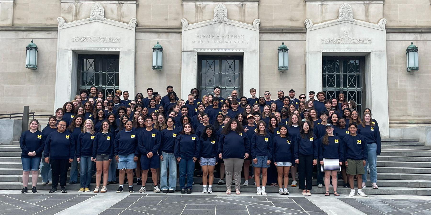 A large group of students wearing matching navy blue Michigan sweatshirts poses for a photo on the steps of a stone building labeled "Horace H. Rackham School of Graduate Studies.