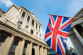Union Jack flag in front of historic stone buildings with columns, photographed from a low angle under a blue sky.