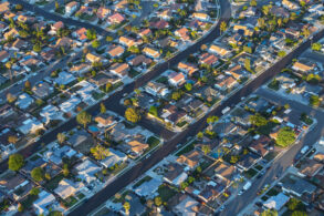 Aerial view of a suburban neighborhood with rows of houses, streets, driveways, and scattered trees in a grid-like pattern.