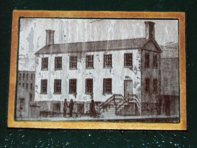 An old, black-and-white illustration of a two-story rectangular building with multiple windows, a front staircase, and a few people standing outside.