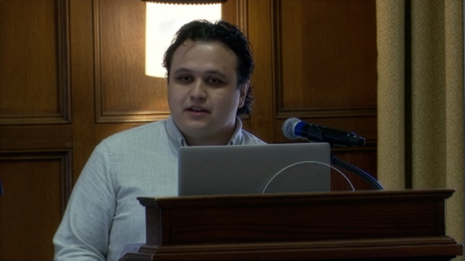 A person stands at a podium with a laptop and a microphone, speaking in a wood-paneled room with beige curtains.