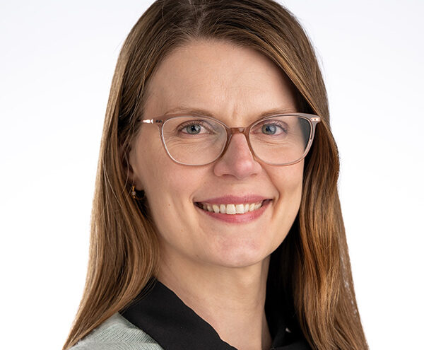 A woman with long brown hair, wearing glasses and a light grey top with a black collar, smiles at the camera against a plain white background.
