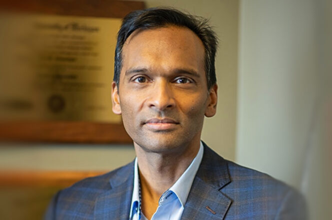 A man in a checked suit jacket and light blue shirt stands indoors, with a blurred framed certificate or diploma in the background.