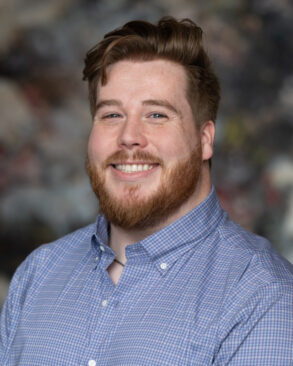 A man with red hair and a beard, wearing a blue checkered button-up shirt, smiles at the camera with a blurred background.