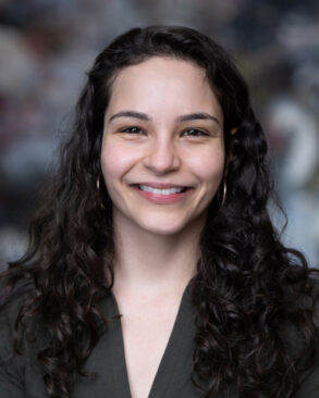 A woman with long, curly dark hair and hoop earrings smiles at the camera, wearing a dark V-neck top. The background is blurred.