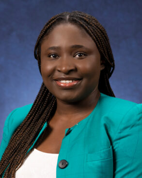 A woman with long braided hair, wearing a teal blazer and white top, smiles in a professional portrait against a dark blue background.