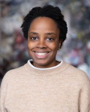 A woman with short, natural hair smiles at the camera, wearing a beige sweater against a softly blurred, multicolored background.